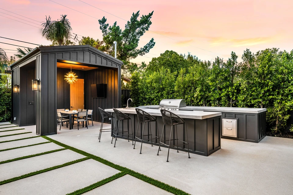Une cuisine extérieure moderne et un coin repas avec un élégant mobilier noir et une élégante pergola, le tout dans un jardin verdoyant au coucher du soleil